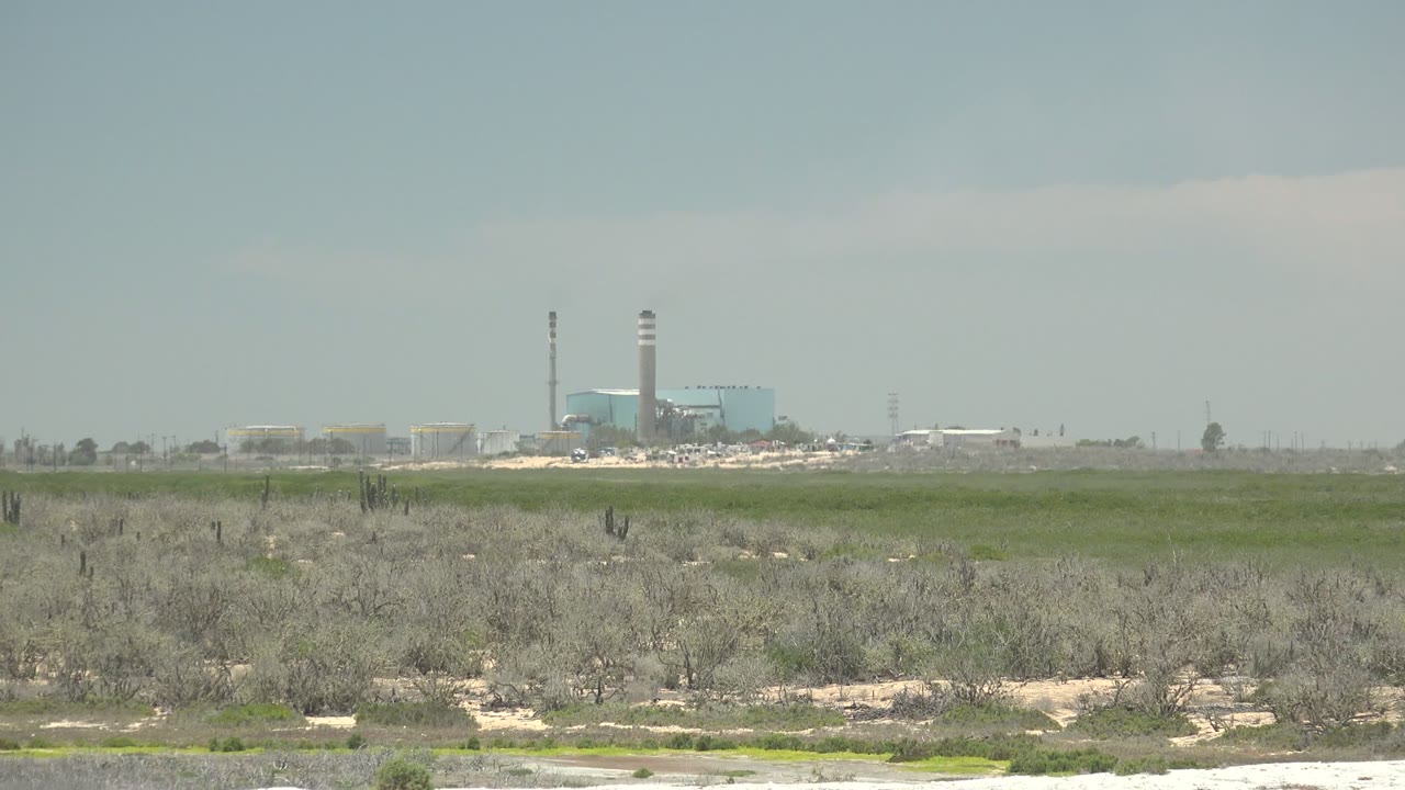 A wide San Carlos, Baja California Sur frame shows low desert vegetation, a pale sandy strip near the bottom, and distant industrial buildings with a tall smokestack beneath light clouds, with no visible people.