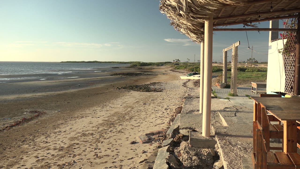 San Carlos, Baja California Sur beach view with low waves, sandy shore, rock edging, patio posts, a thatched roof, and no visible people.