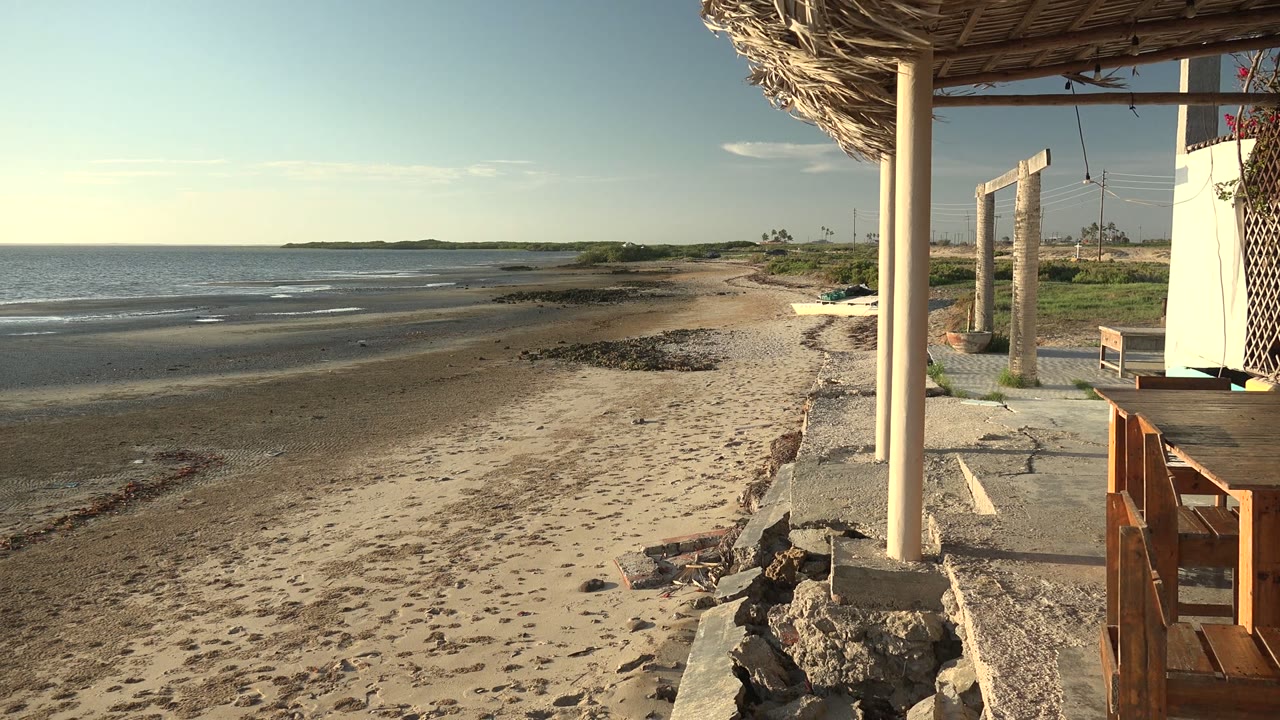 San Carlos, Baja California Sur shoreline framed by a thatched shelter, with wooden furniture at right, rippled water at left, and no visible people.
