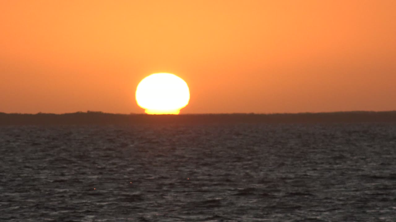 At 0:00, the sun sits just above the distant land horizon over dark ocean water in San Carlos, Baja California Sur, with orange sky and no visible people.