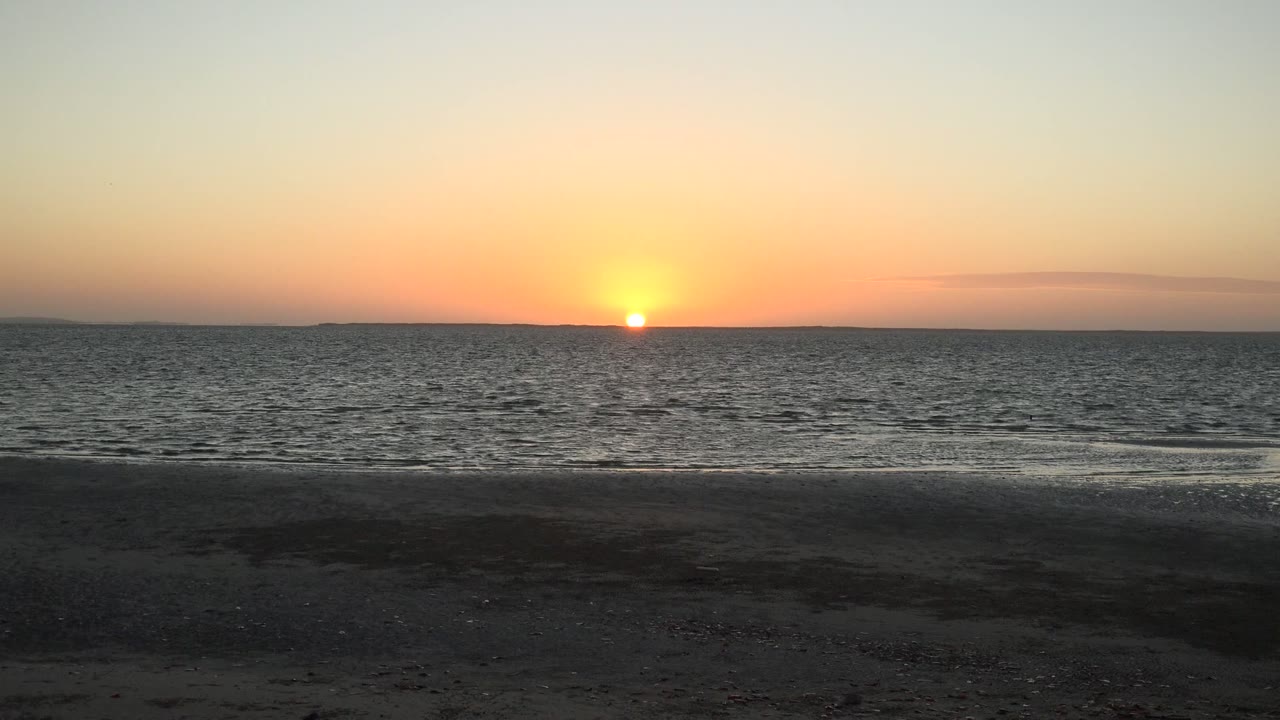 Sunset over the ocean at San Carlos, Baja California Sur, with rippled water, dark sand in the foreground, warm orange sky, and no visible people.