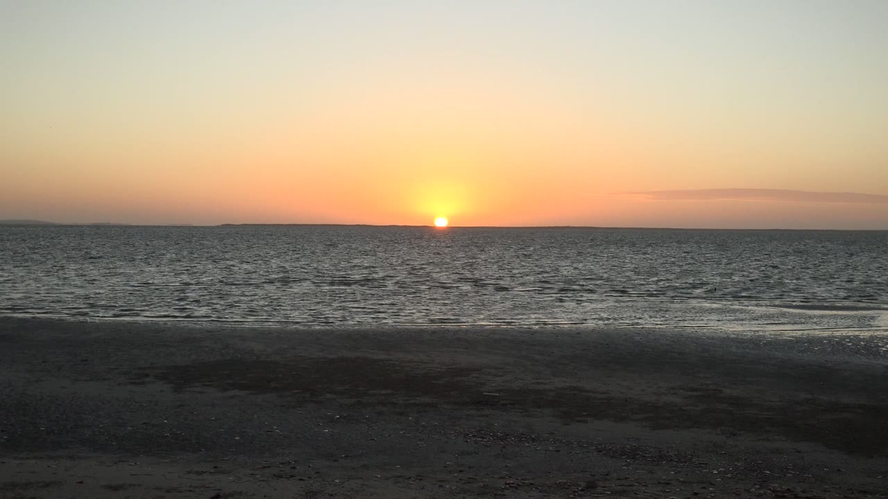 Low sun on the horizon above calm ocean and sandy shore.