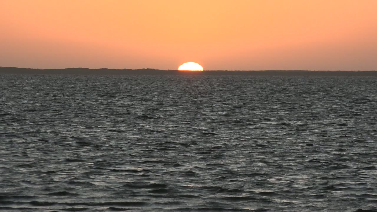In San Carlos, Baja California Sur, Mexico, the half-set sun touches the dark distant horizon above rippled ocean water, with no visible people.