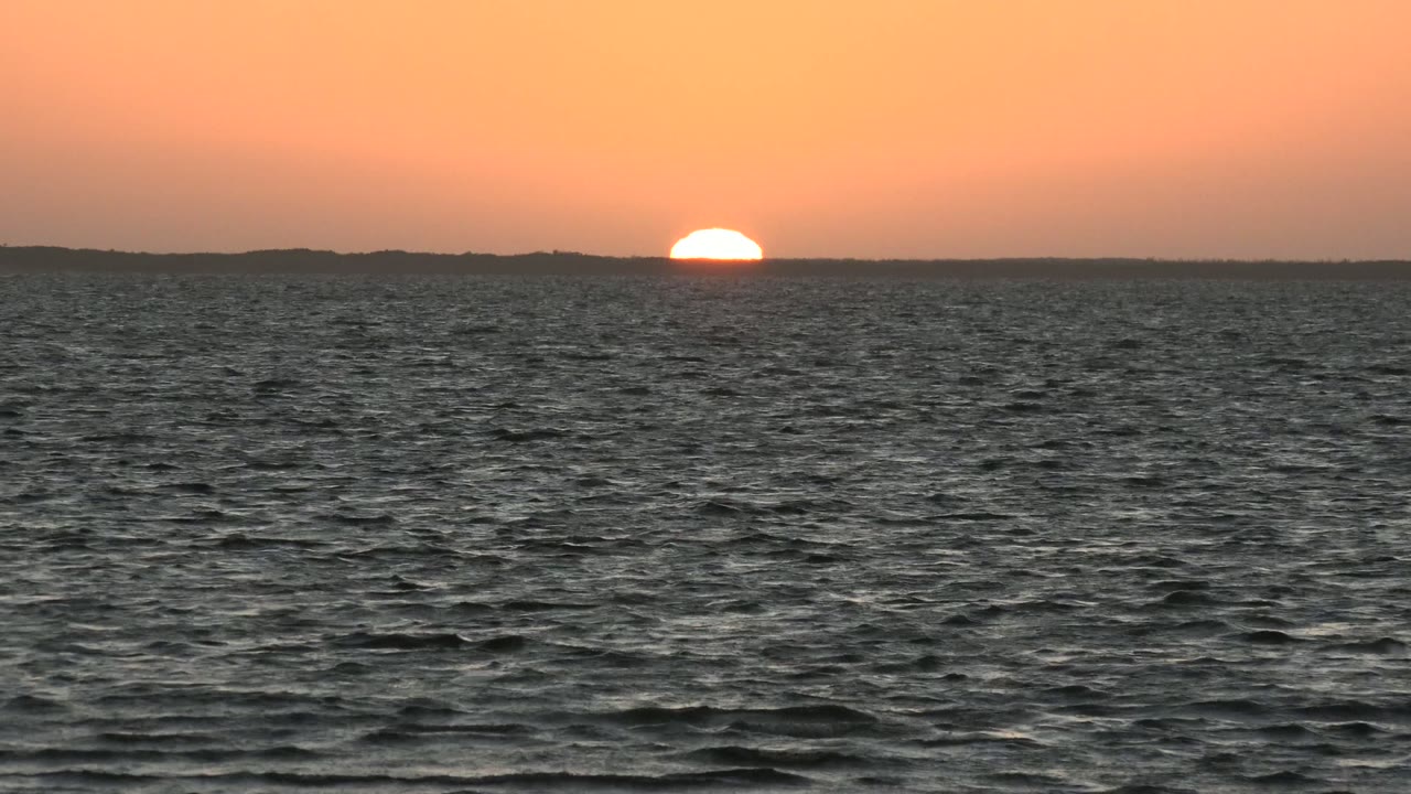 A bright half-disc of sun rests on the horizon beyond the open sea, with distant land silhouetted below an orange sky and no visible people.