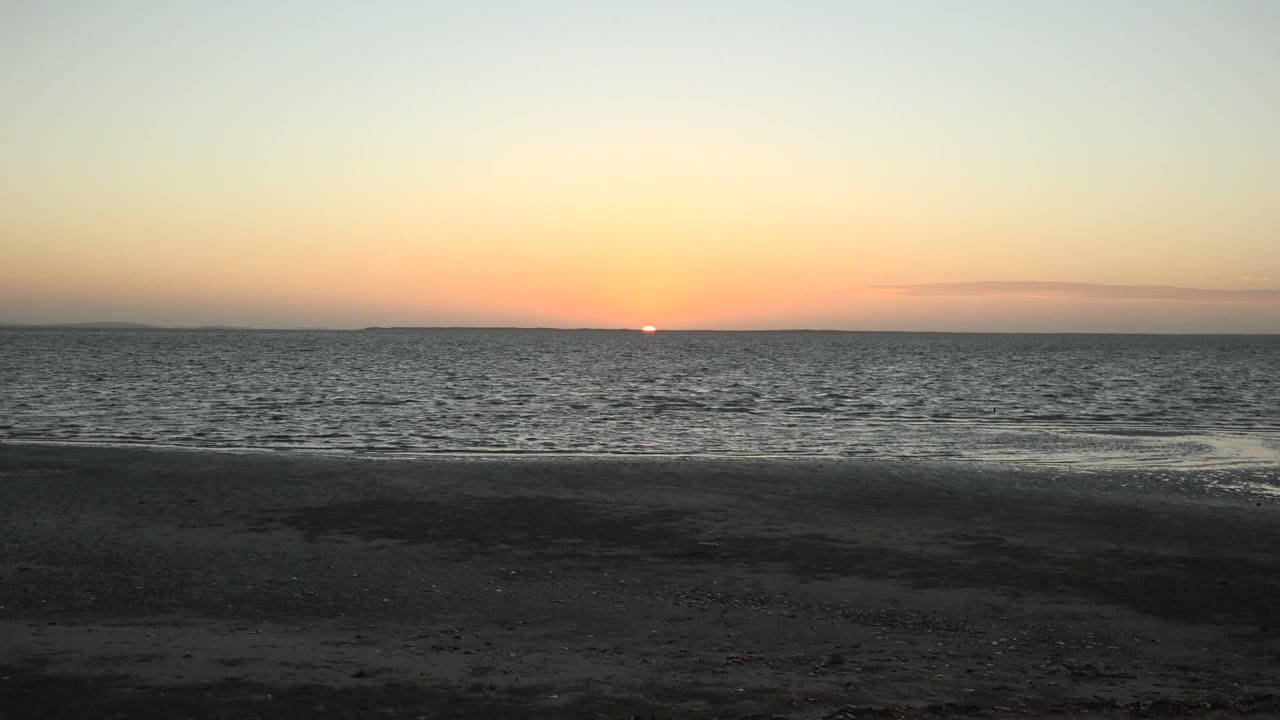 Opening frame of a San Carlos, Baja California Sur sunset with the sun just above the horizon, rippled water in the middle distance, dark sand in front, and no visible people.