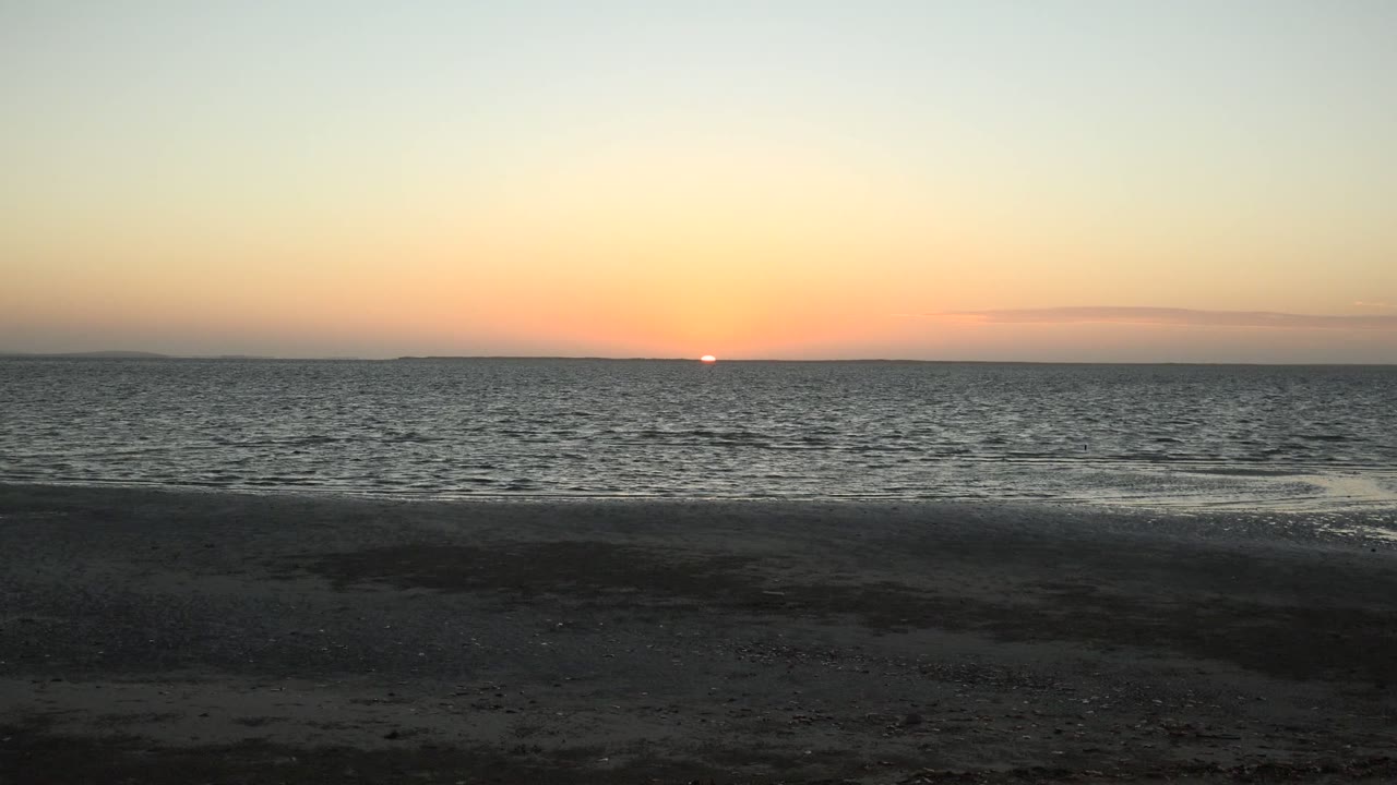 At 0:01, the San Carlos coastal view holds the sun near the horizon while a thin wave line brightens the edge of the dark sandy beach, with no visible people.