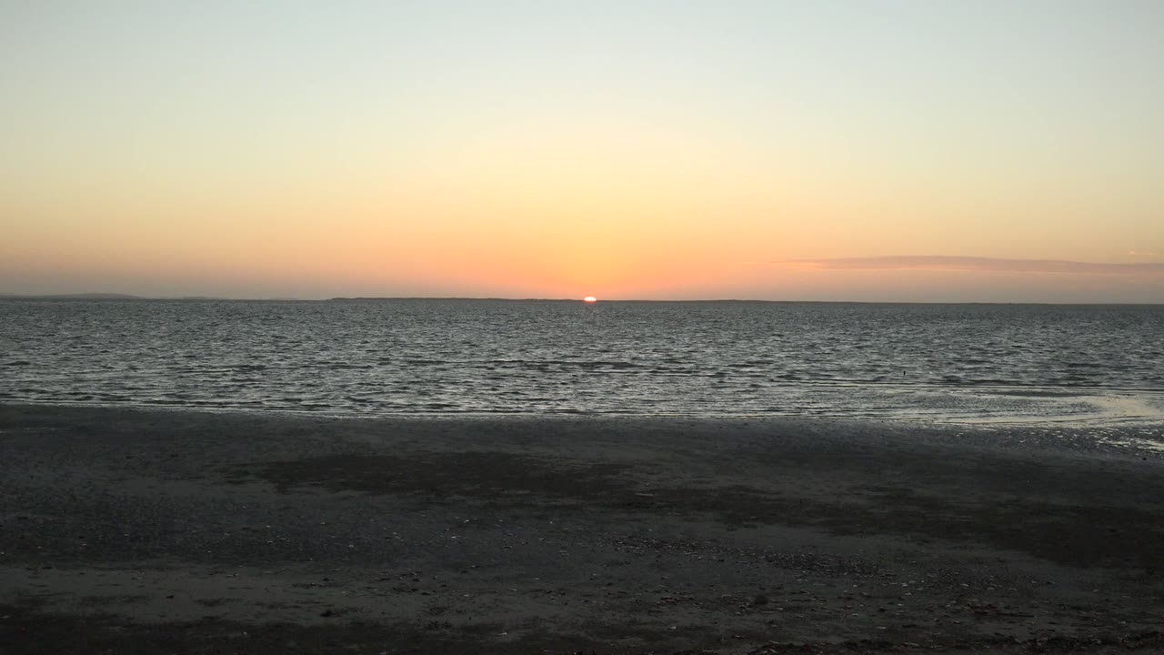 At 0:02, the sun sits low over the San Carlos ocean horizon as a pale wash of water spreads near the right side of the sandy foreground, with no visible people.