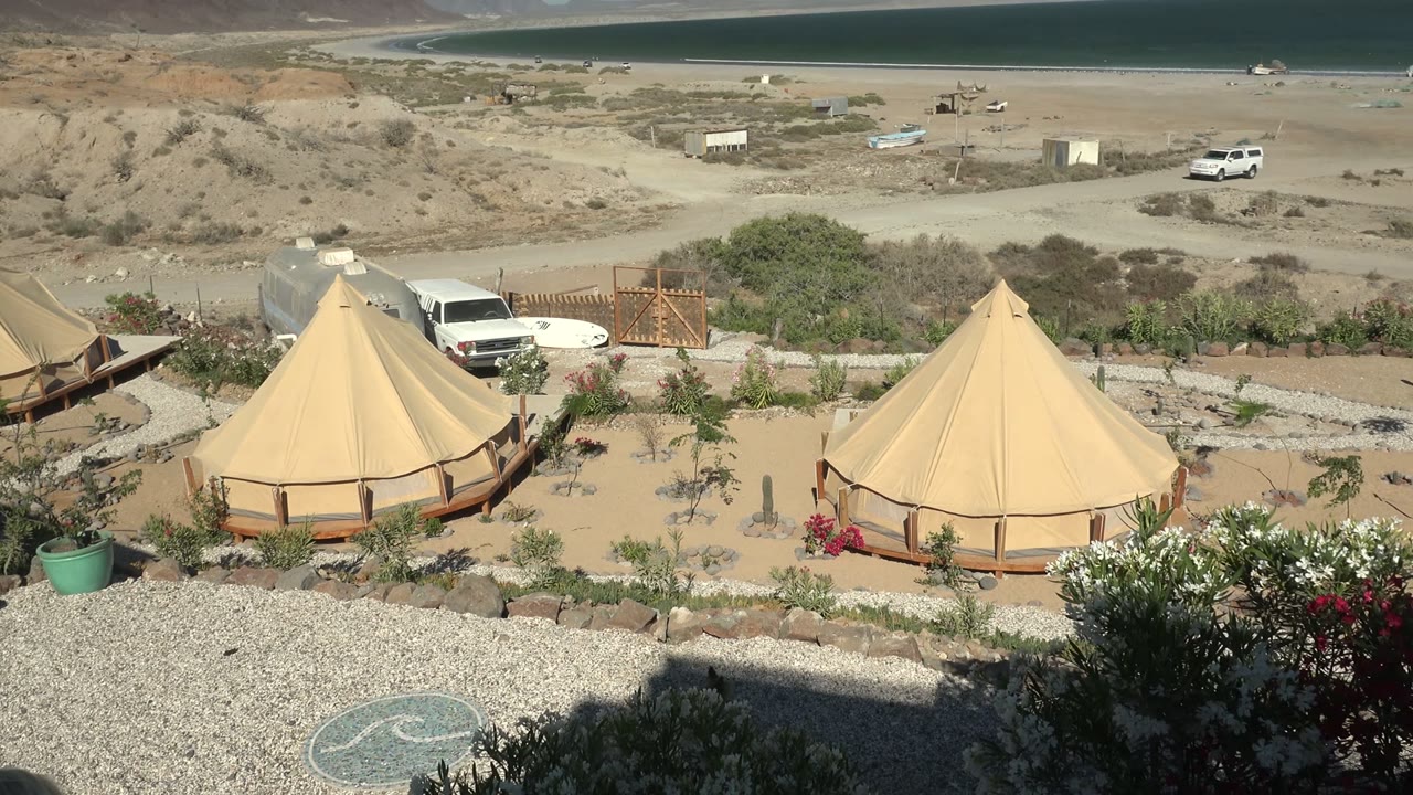 Opening frame of tan bell tents on sandy ground in San Juanico, Baja California Sur, with parked vehicles, scrubland, distant water, and no visible people.