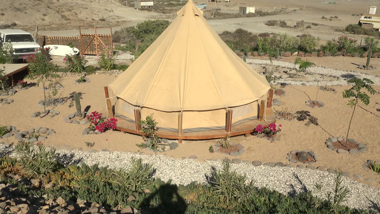 A beige round tent in San Juanico, Baja California Sur sits amid gravel, desert plants, red flowers, parked vehicles, distant hills, blue ocean, and no visible people.