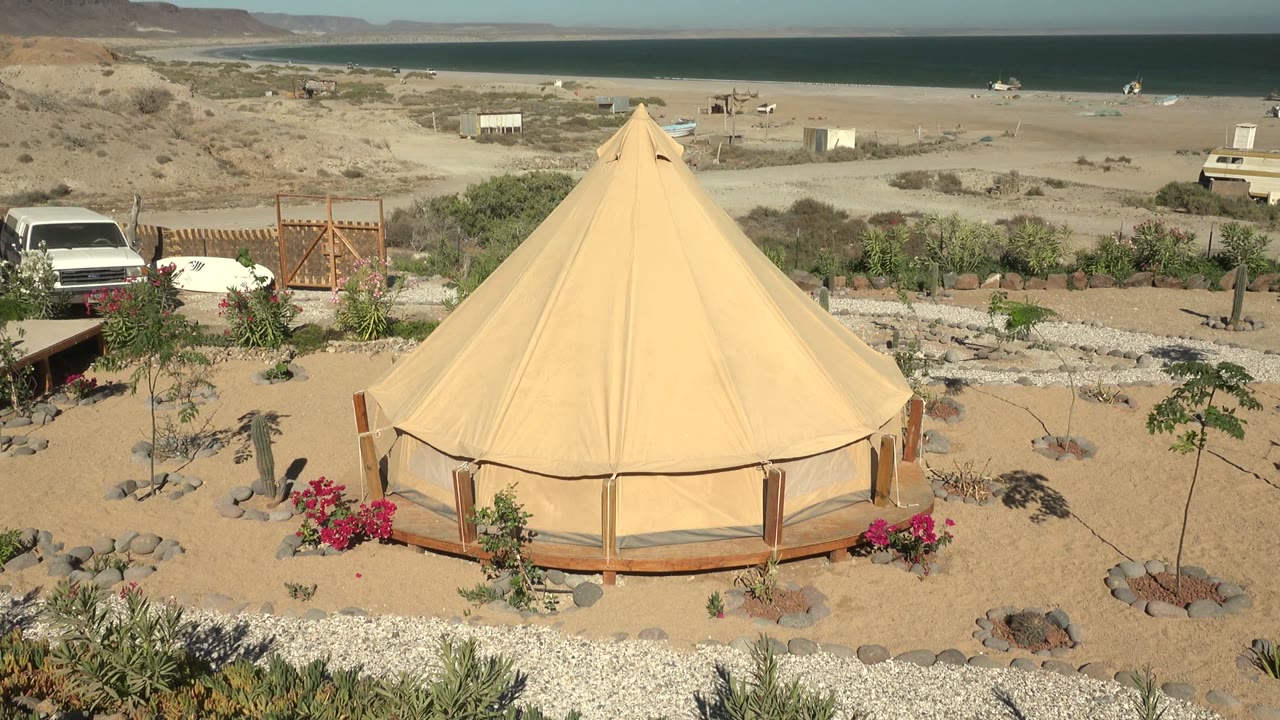 A beige yurt-like tent in San Juanico fills the foreground with desert plantings and rocks, while the ocean horizon, low hills, vehicles, and no visible people appear behind it.