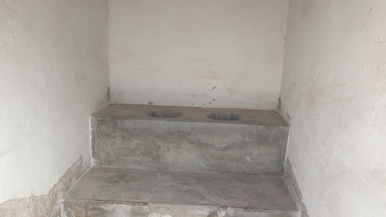 Early view in Mulege, Baja California Sur, Mexico, showing a gray concrete squat toilet platform with two circular openings, worn white walls, and no visible people.