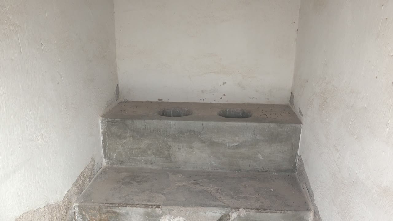 The frame shows the unfinished Mulege room corner with gray concrete steps below a squat toilet platform, two circular openings, worn white walls, and no visible people.