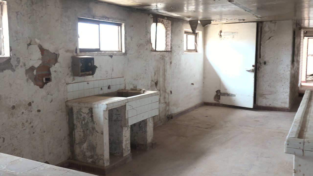 Opening frame of a dilapidated washroom in Mulege, Baja California Sur with a stained tiled sink at left, peeling white walls, small windows, a bright door, and no visible people.
