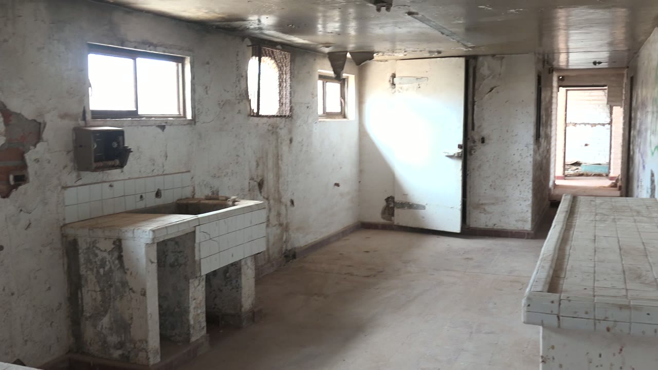 Abandoned washroom corner with tiled sink, cracked wall plaster, windows, and door.