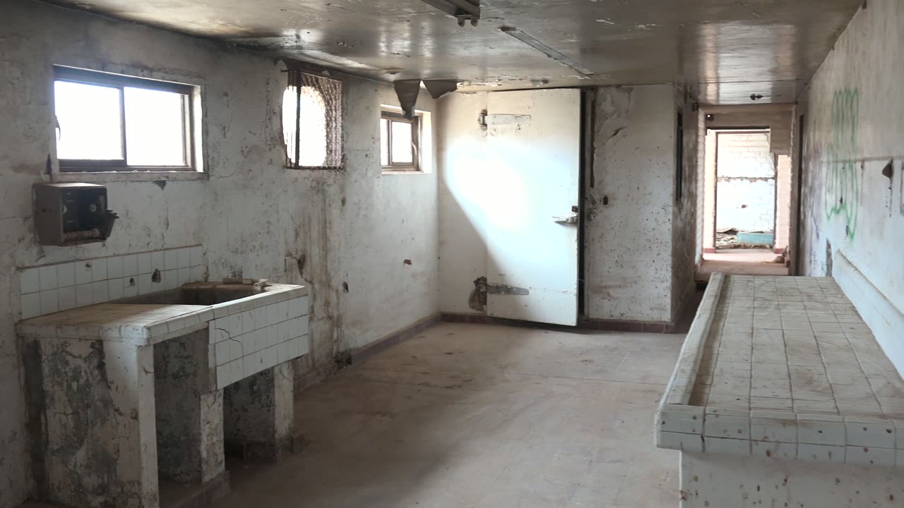 A long tiled counter runs from the foreground toward the back doorway in the Mulege washroom, with dirty walls, windows, and no visible people.