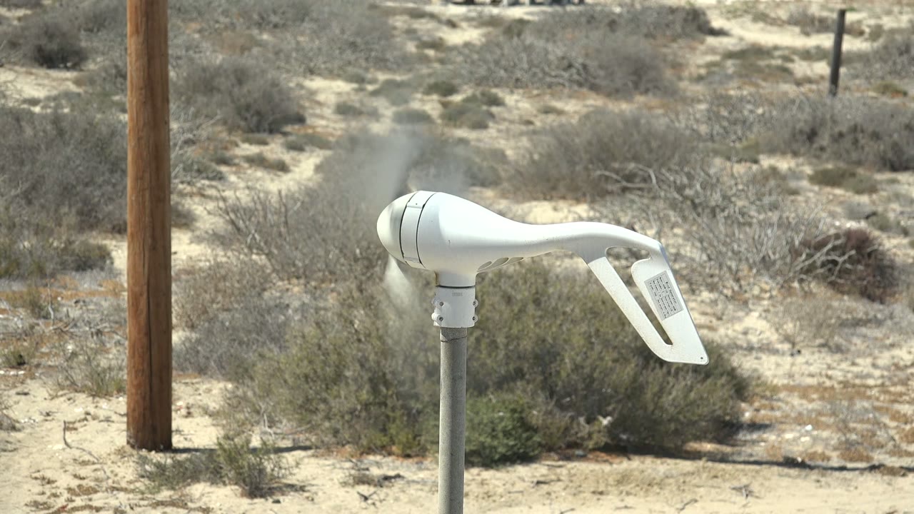 White wind vane spins on a metal pole in La Bocana, Baja California Sur, with sandy ground, dry shrubs, a wooden utility pole, and no visible people.