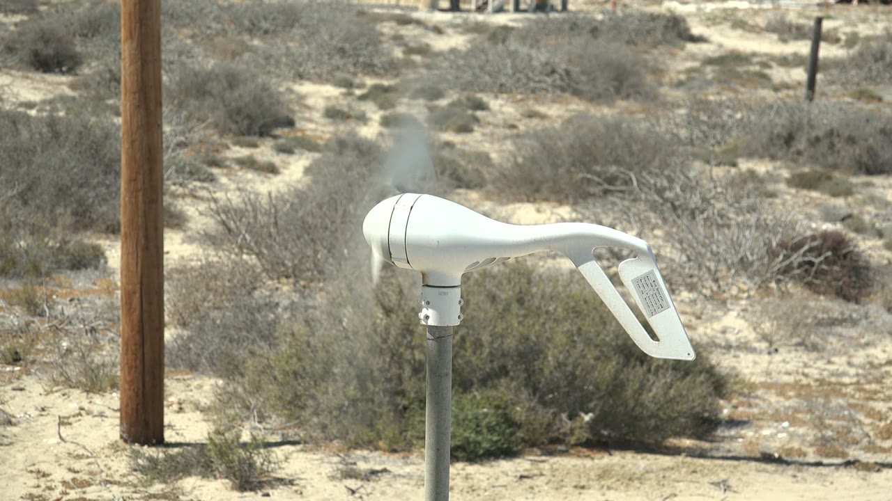 A white wind vane remains centered on its pole in La Bocana, Baja California Sur, angled over sandy brushland with a wooden utility pole at left and no visible people.