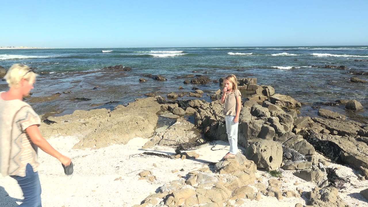 At La Bocana, Mexico, an adult enters from the left while a child stands on shoreline rocks with shallow ocean and white wave lines behind them.