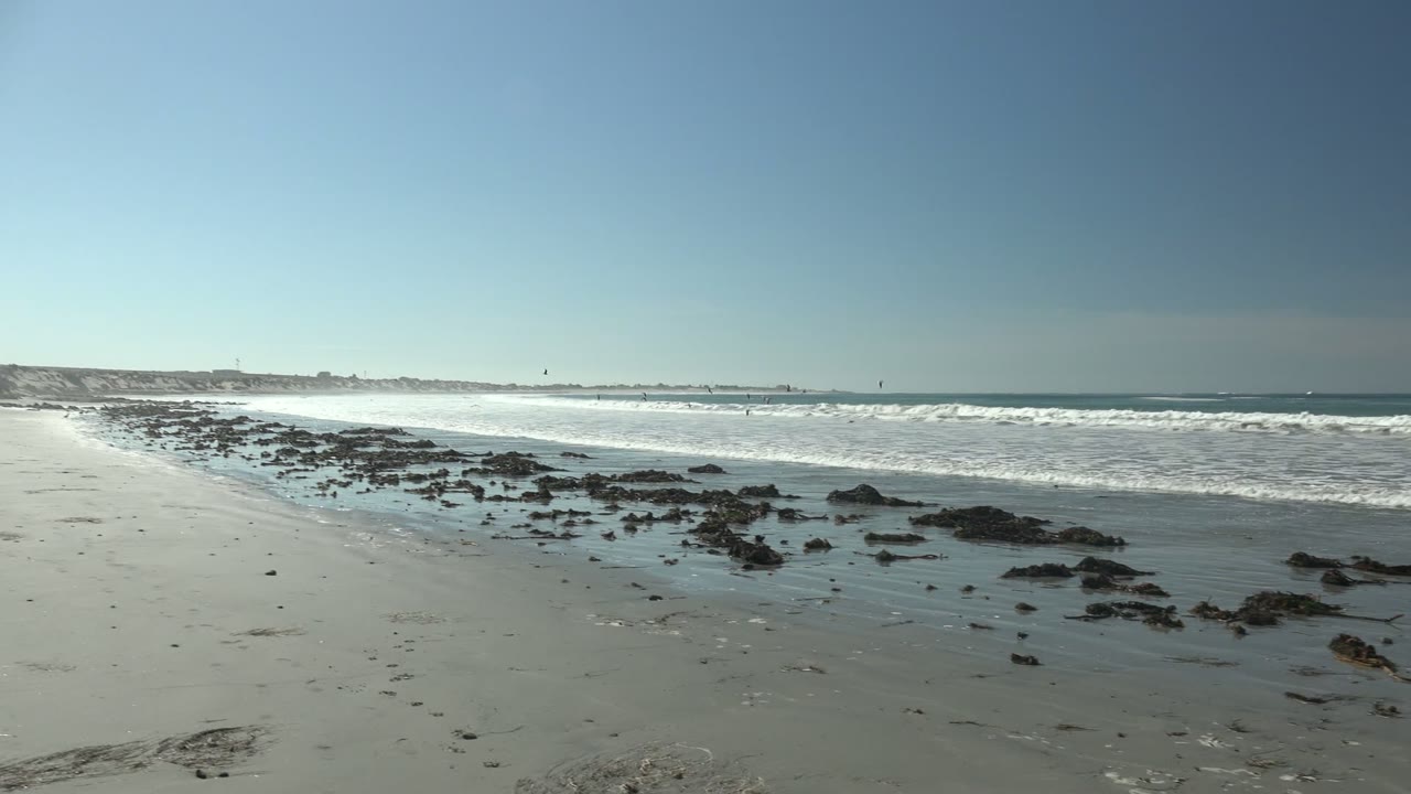 Wide view of La Bocana beach with wet sand, scattered dark rocks, white surf bands, clear blue sky, and distant water activity near the horizon.