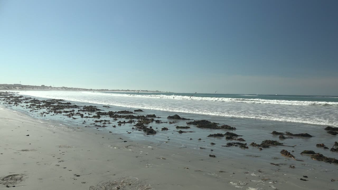 La Bocana shoreline with low rocks spread across reflective wet sand, white waves breaking behind them, and small distant figures or kites offshore.