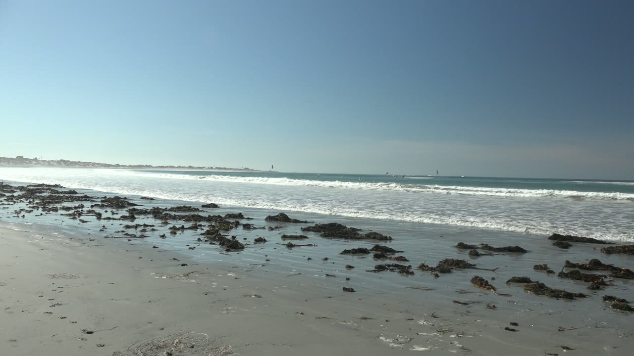 Low beach rocks and shallow water sit in the foreground at La Bocana while a wide sheet of surf rolls toward shore beneath a clear sky.