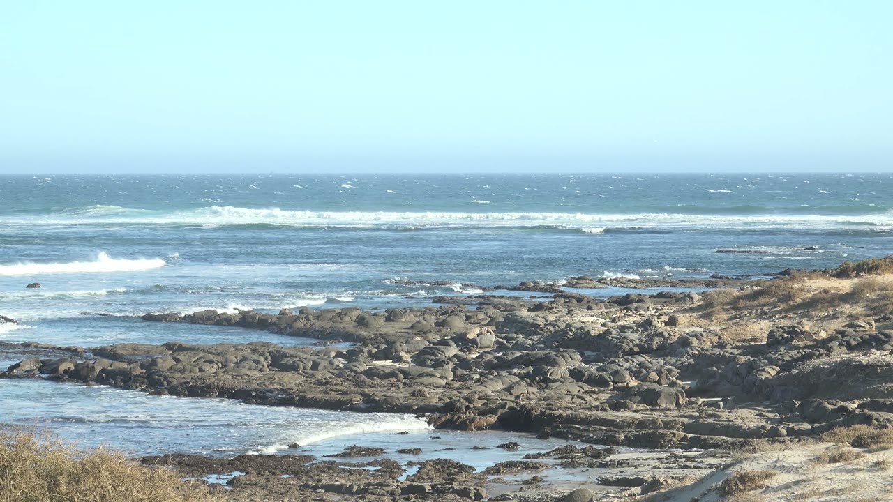 At La Bocana, Baja California Sur, waves break beyond dark shoreline rocks and shallow tide pools under a pale sky, with no visible people.