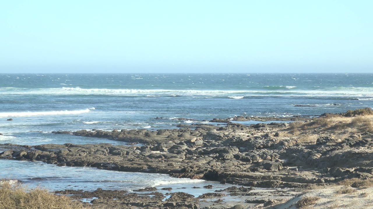 Whitecaps form across the open water at La Bocana while uneven rocks and reflective tide pools fill the lower frame, with no visible people.