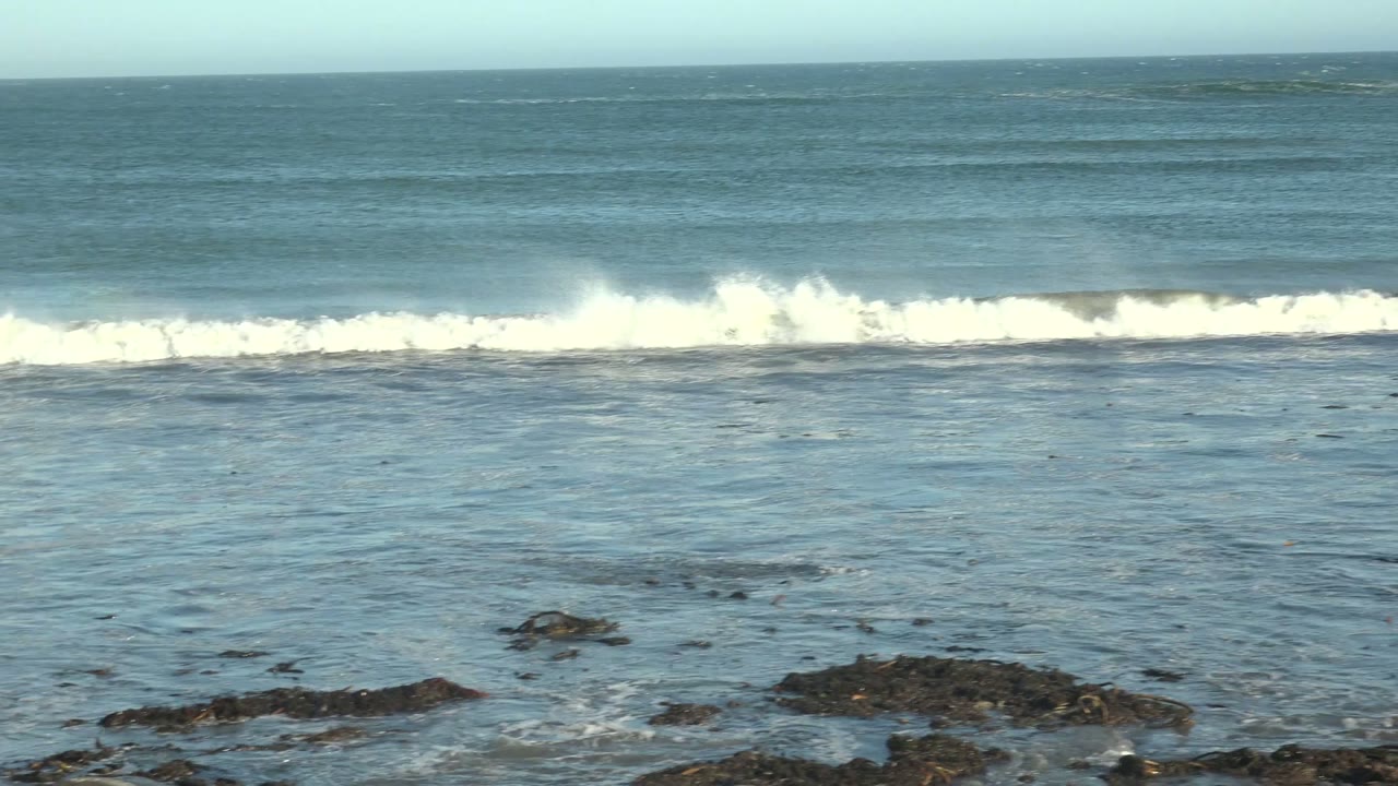 White foam spreads along the midground surf line at La Bocana while shallow water covers dark rocks in the foreground, with no visible people.