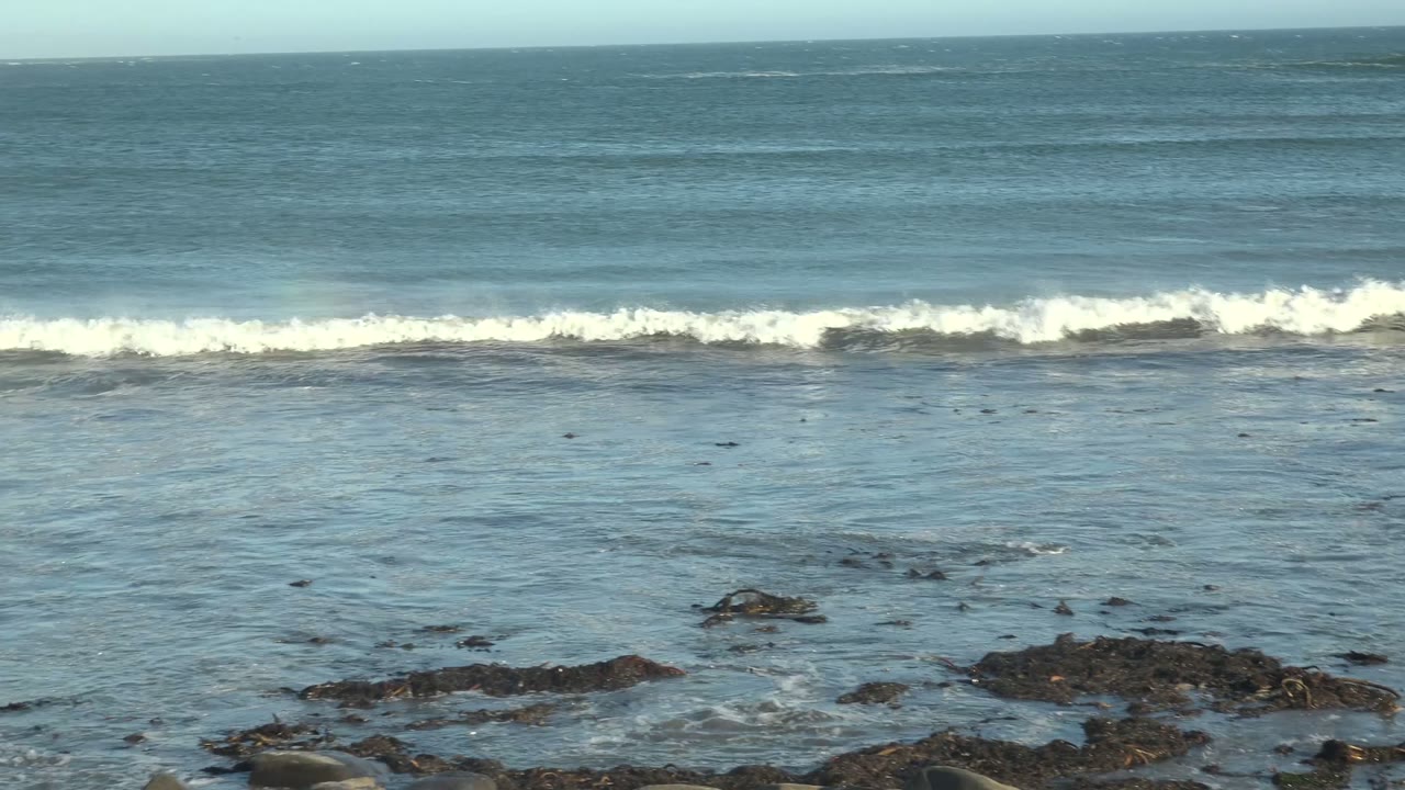 The broken wave pushes shoreward as white foam crosses the lower midframe, with La Bocana's rocky foreground exposed beneath the shallow wash and no visible people.