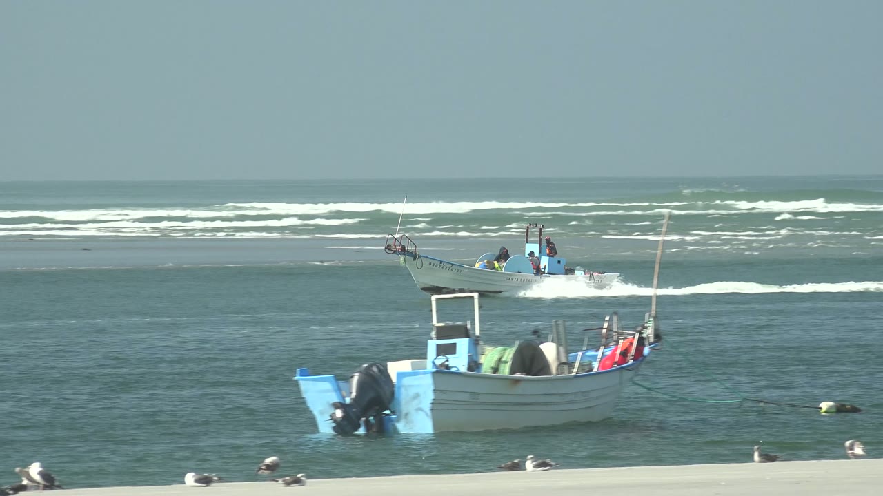 Two small boats sit and move on green-blue water off La Bocana, Baja California Sur, with seagulls on the pale sand and white surf bands in the distance, no visible people identifiable.