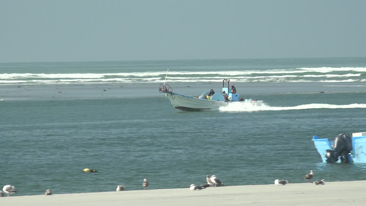 A small boat travels across open coastal water in front of white surf at La Bocana, Baja California Sur, while another blue and white boat rests closer to the sandy beach.