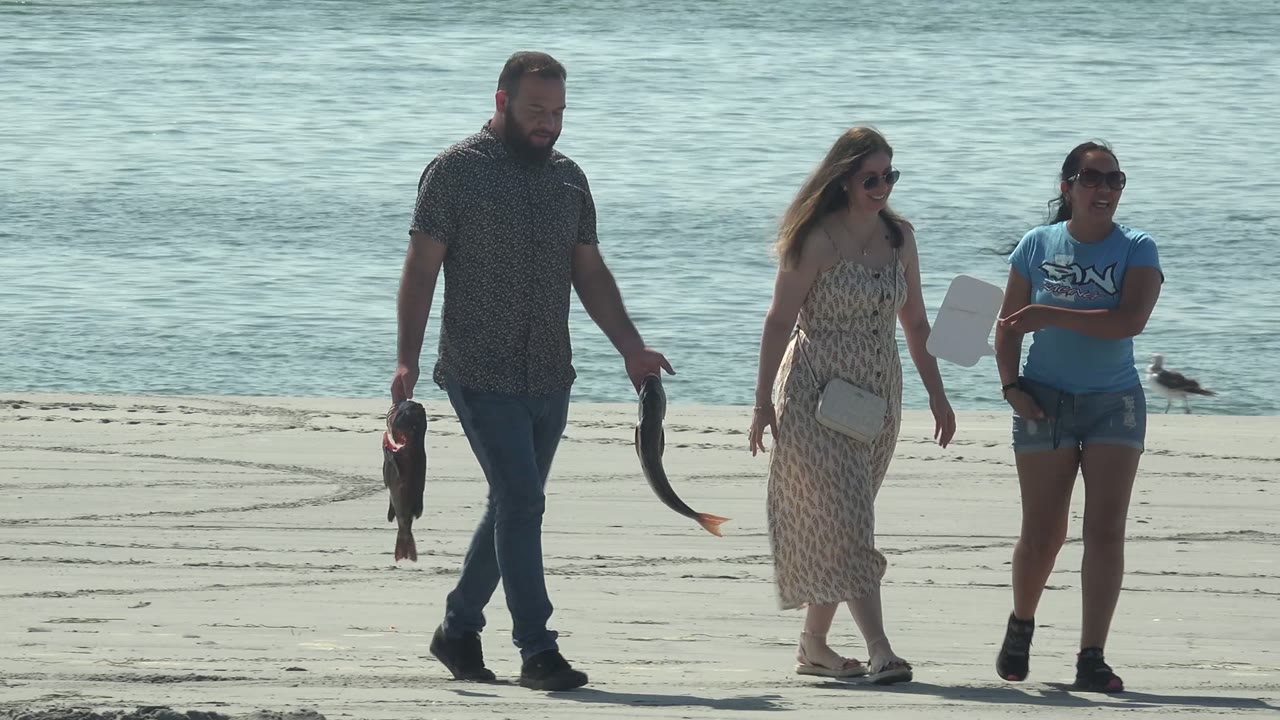 Three adults walk along the sandy shore in La Bocana, Baja California Sur, with the man at left holding two fish and blue-green water behind them.