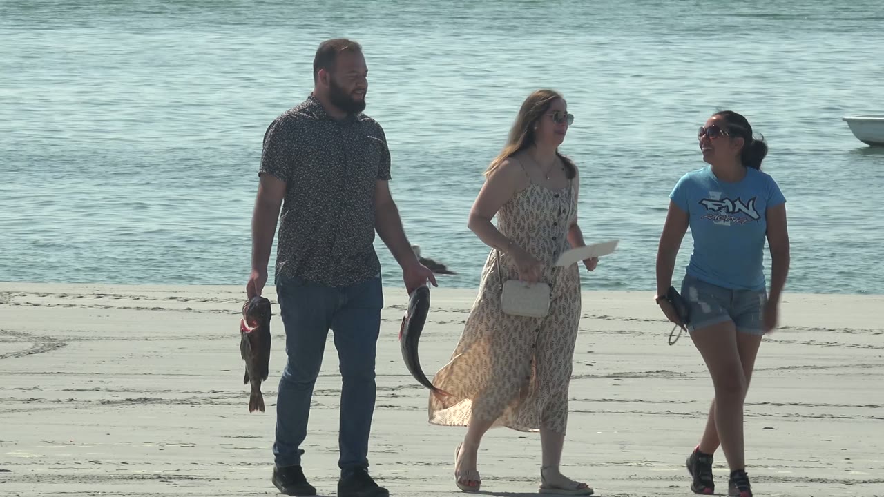 Three adults move farther across the sandy La Bocana beach, the man still holding two fish while the women walk beside him near the waterline.
