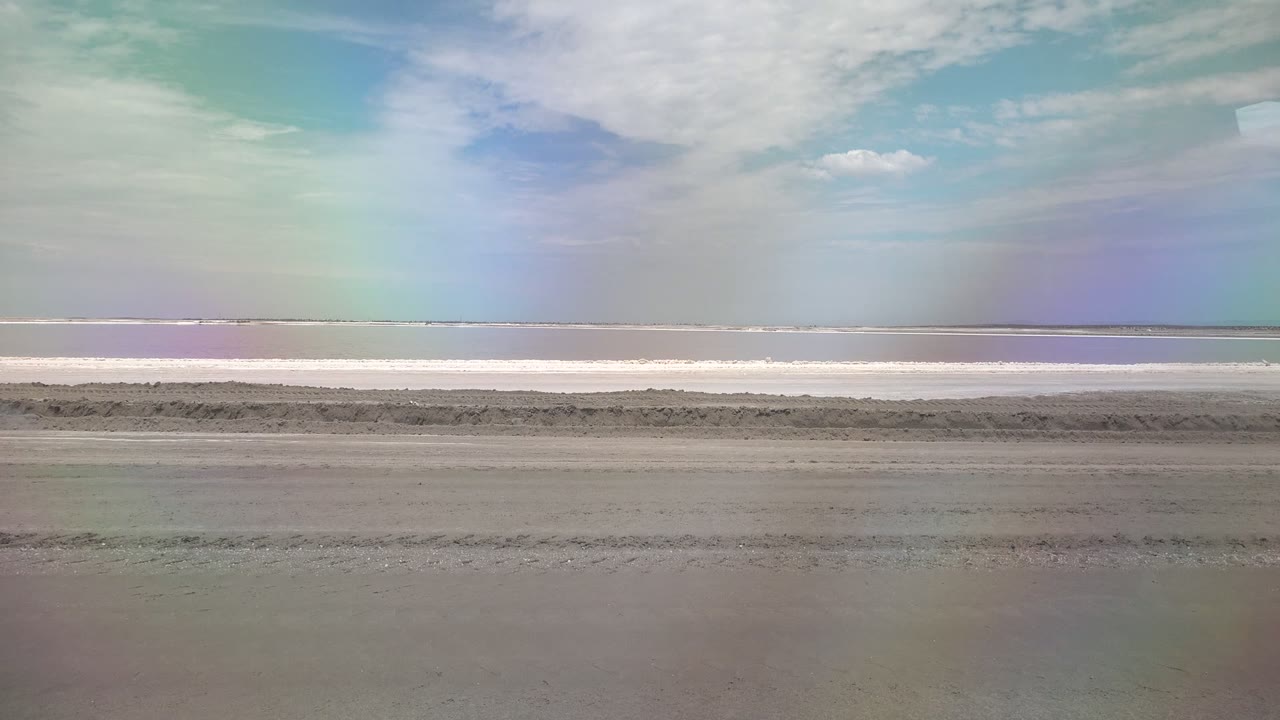 Wide Guerrero Negro beach frame with wet sand tracks, a pale waterline, cloudy blue sky, rainbow color across the sky, and no visible people.