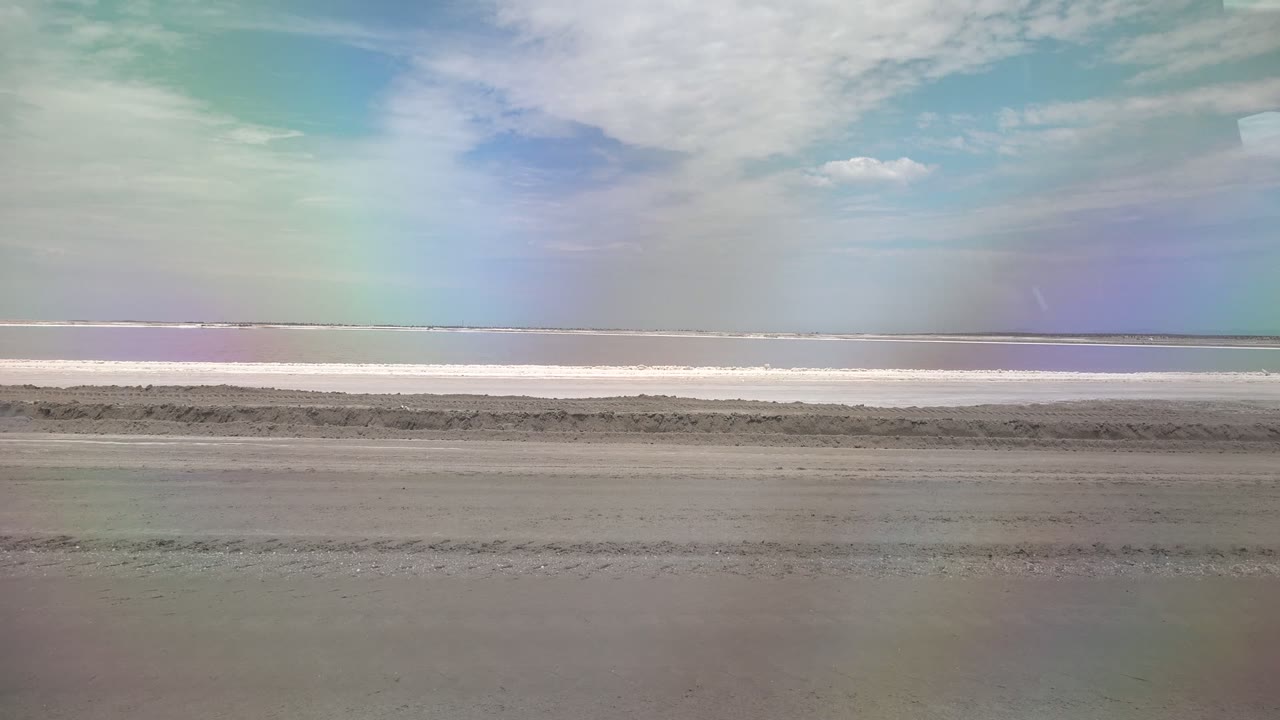 Empty shoreline with wet sand, waterline, clouds, and rainbow tint in the sky.