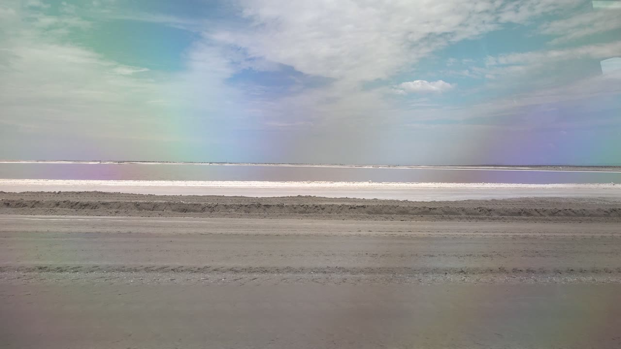 A wide Guerrero Negro beach view at 0:02 shows wet sand foreground texture, a thin distant waterline, soft clouds, rainbow color in the sky, and no visible people.