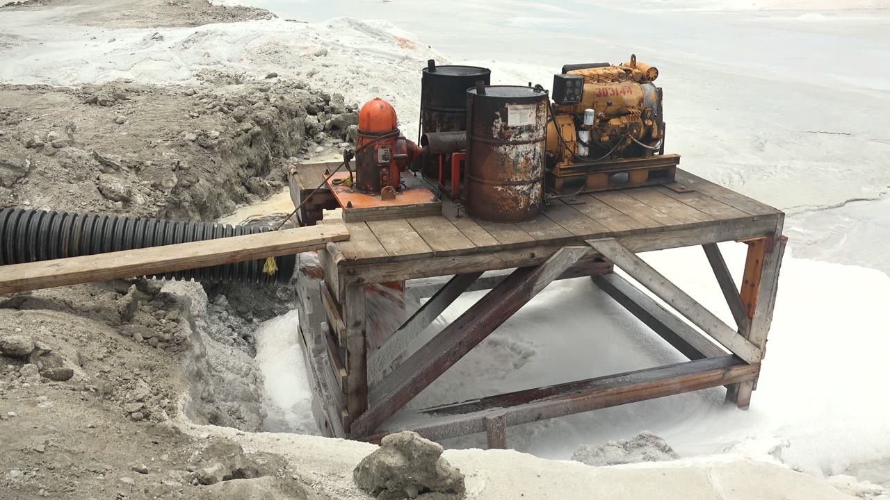 At 0:01, the pump station remains centered on its wooden platform, with rough gray earth at left and white frozen surface behind it in Guerrero Negro, Mexico, with no visible people.