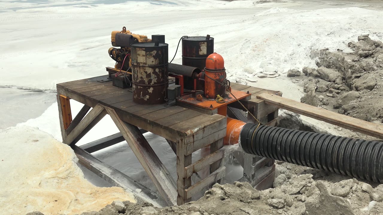 Rusted pump machinery on a wooden platform connects to a large black ribbed hose over pale slurry and muddy ground in Guerrero Negro, Mexico, with no visible people.