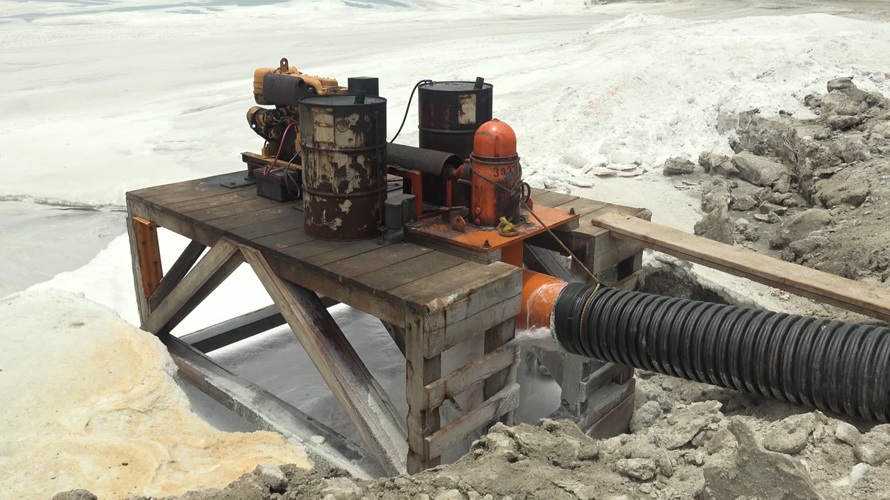 A rust-stained cylinder, orange tank, and black hose sit on the platform above pale slurry channels and gray mud in Guerrero Negro, Mexico, with no visible people.