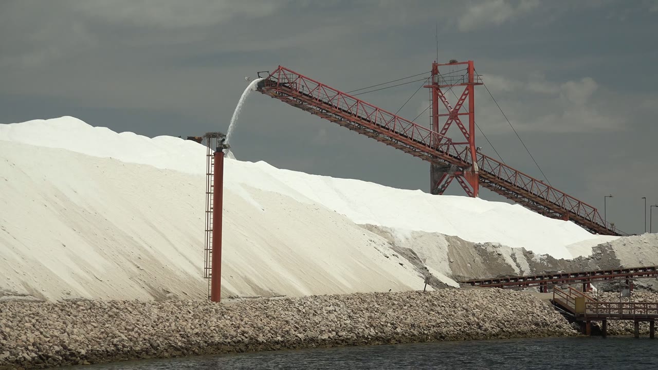 At 0:00 in Guerrero Negro, Baja California Sur, white salt piles sit behind shoreline rocks while a red conveyor and spray tower cross the frame, with no visible people.