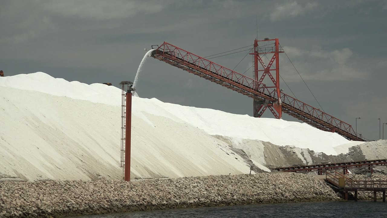 At 0:02 in Guerrero Negro, Baja California Sur, the red conveyor spans diagonally across the white salt piles as the spray tower sends water over the mound, with no visible people.