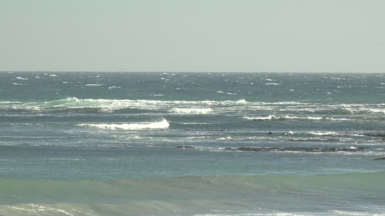 Blue-green water at La Bocana shows white foam bands crossing dark shallow rocks, with a hazy sky above the straight horizon and no visible people.