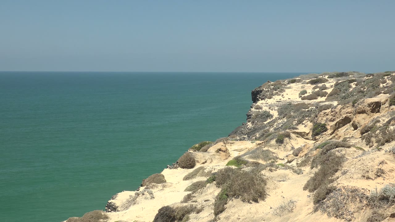 San Quintin BC Mexico cliff view at the start of the clip, with sandy rock and low scrub on the right, turquoise ocean on the left, clear sky above, and no visible people.