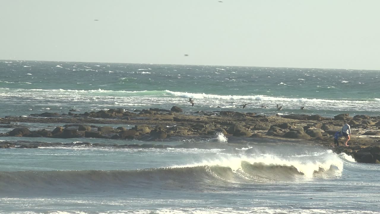 At 0:00 in La Bocana, Baja California Sur, a wide view shows a white wave curling toward dark shoreline rocks, with small distant people on the rocks and birds above the ocean.
