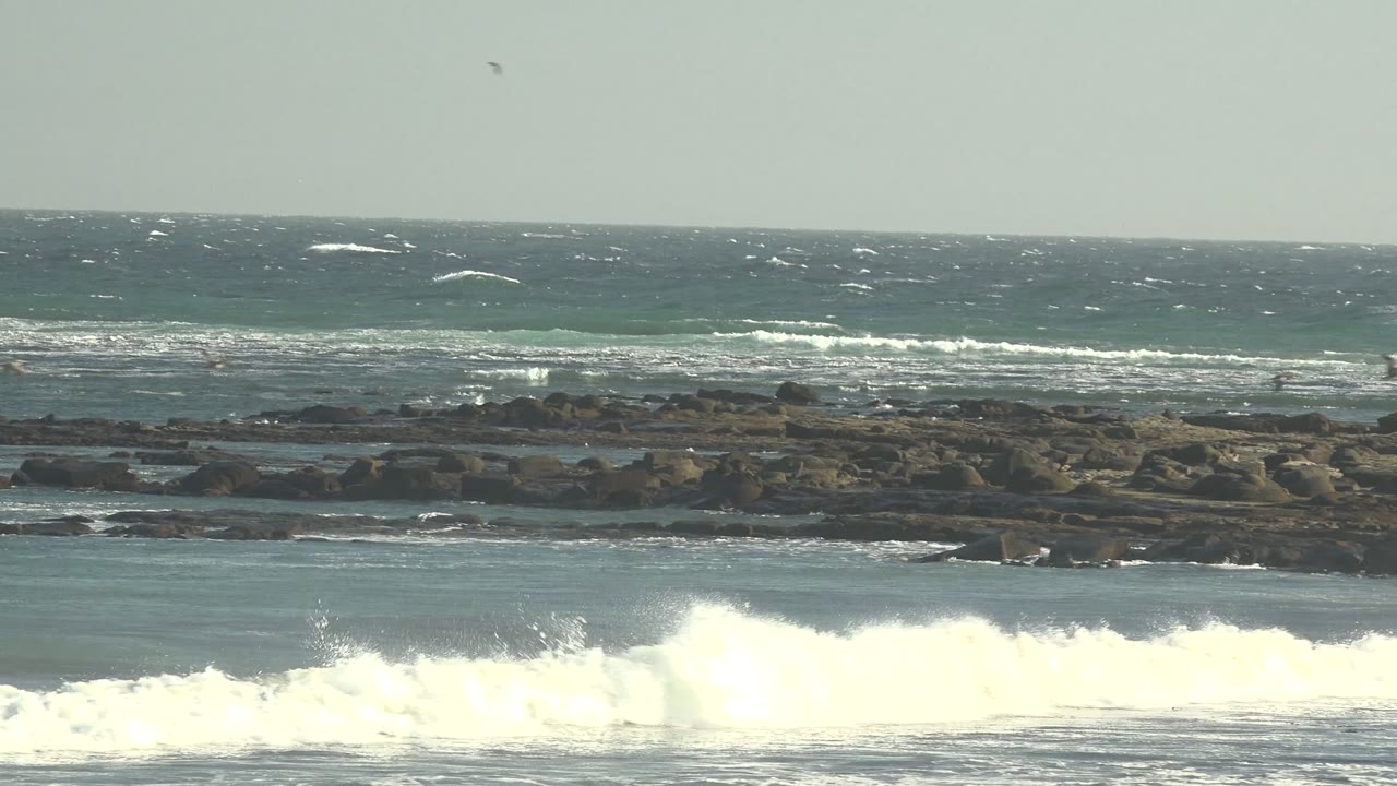 At 0:02 in La Bocana, Baja California Sur, bright whitewater pushes across the right side of the frame while dark rocks, distant people, birds, and a flat ocean horizon remain visible.