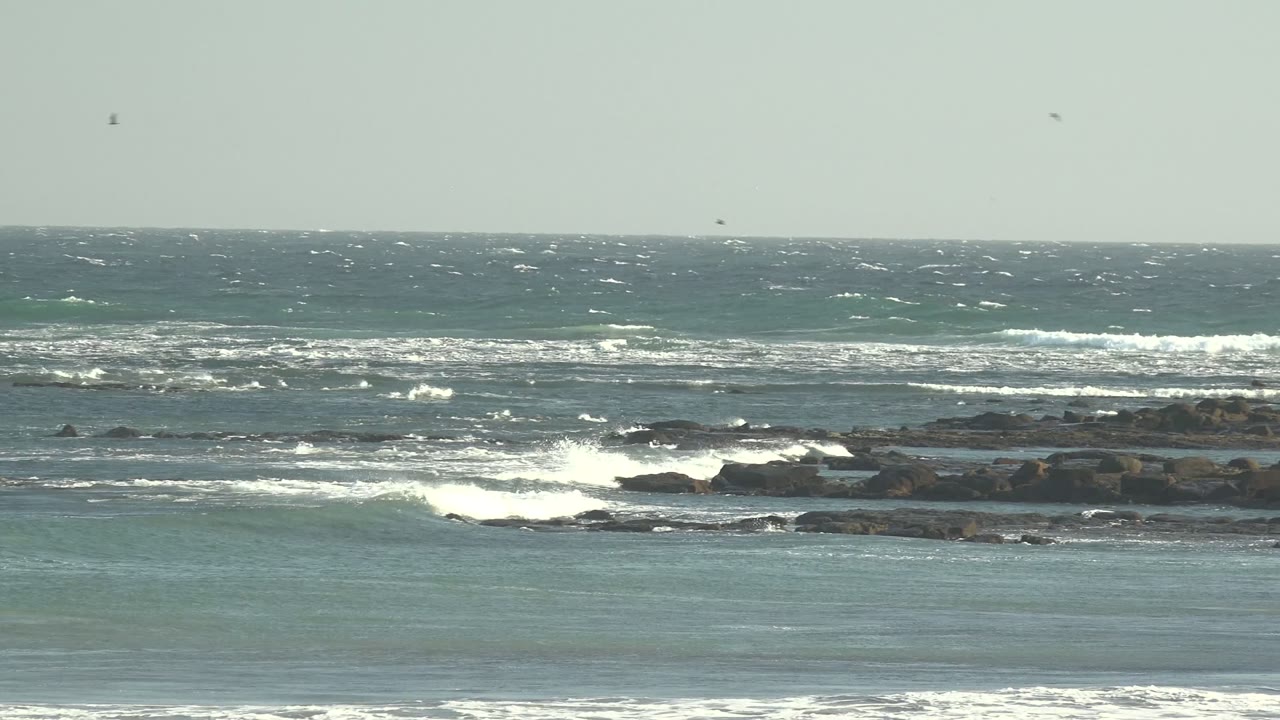 Wide view of blue-green surf breaking over dark rocky shelves at La Bocana, Baja California Sur, with a level horizon and no visible people.
