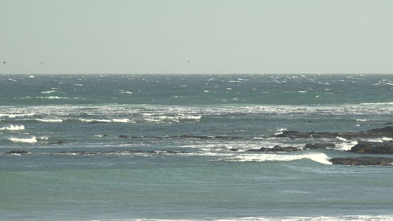Foamy waves break unevenly across dark rock patches at La Bocana, with muted green-blue water and no visible people in the frame.