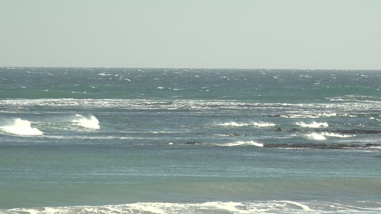 Wide view of blue green ocean surf at La Bocana, Baja California Sur, with whitecaps breaking around dark rocky outcrops and no visible people.