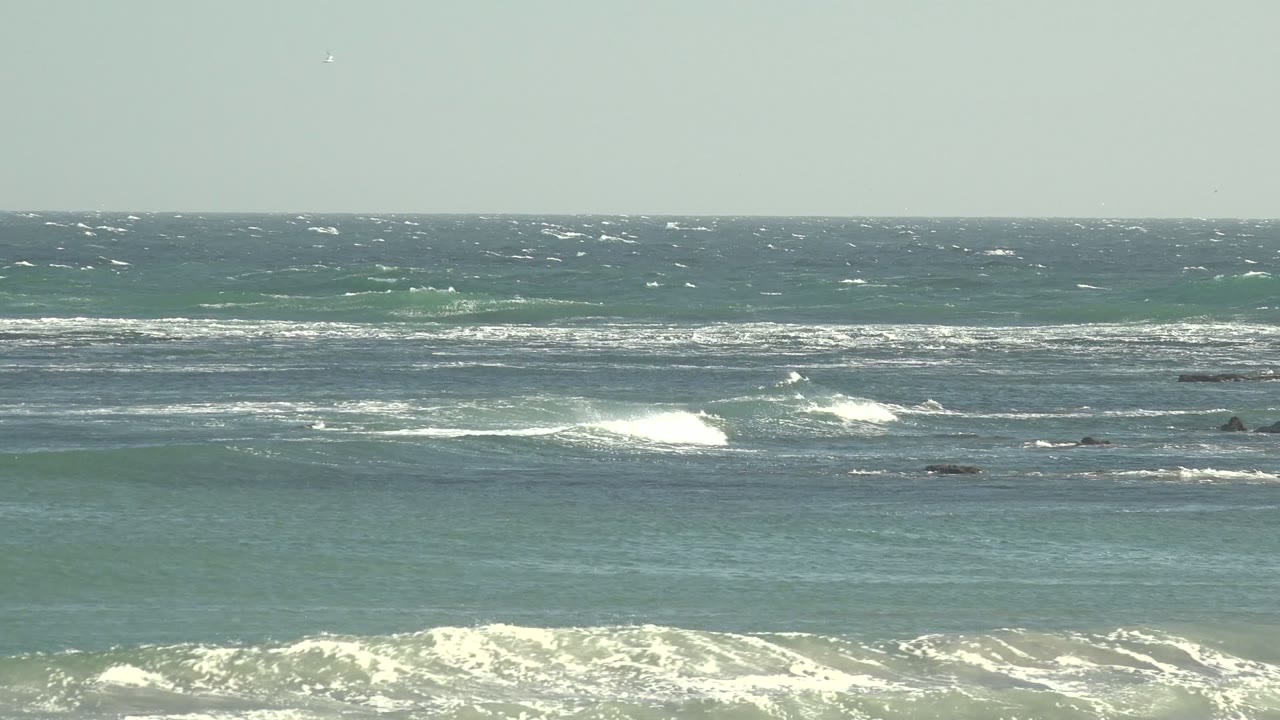 A broad La Bocana surf view shows bright white breaks across blue green water, dark rock patches in the midground, and no visible people.