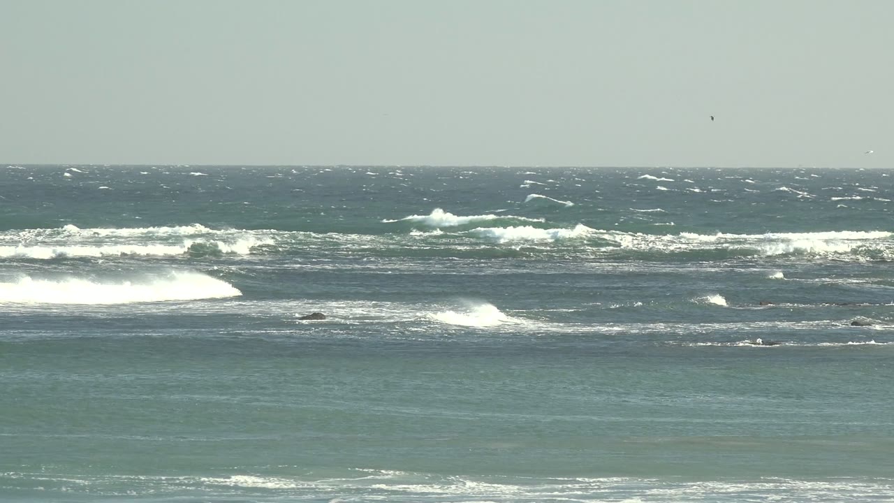 At 0:00 in La Bocana, Baja California Sur, blue-green ocean bands break into whitecaps over a rocky seabed beneath a pale gray sky, with no visible people.